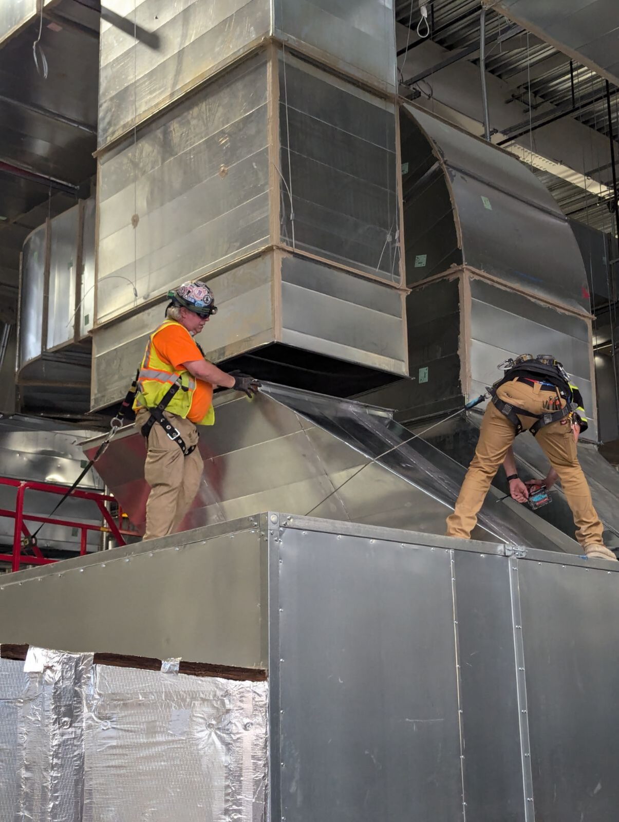 Two union sheet metal workers in harnesses installing large ductwork